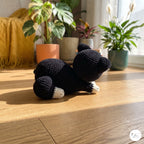 A handmade crocheted black cat toy with white paws sits on a wooden floor in a brightly lit room with potted plants in the background.