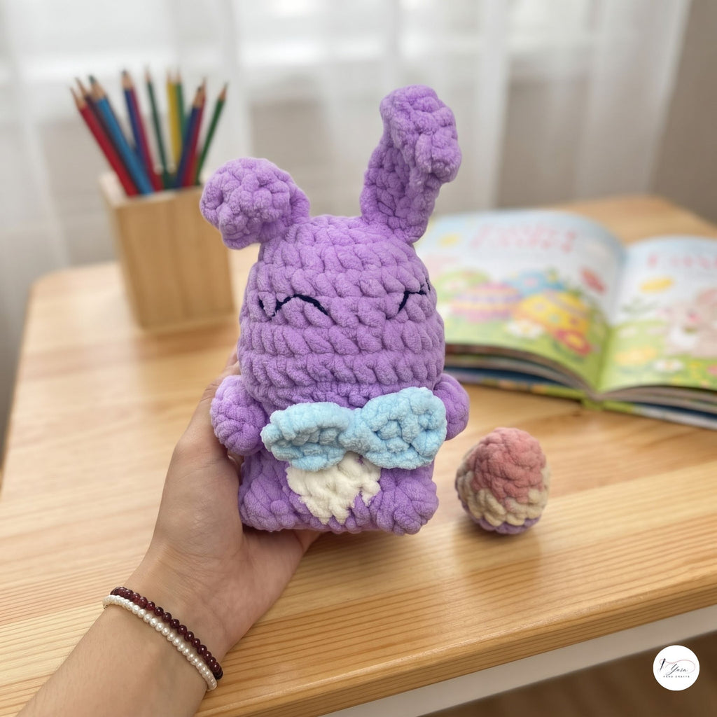 A hand holds a small crocheted purple bunny plush toy with a light blue bow and white belly, with a small matching pink and cream accessory beside it on a wooden table. An open book and pencil holder are blurred in the background.
