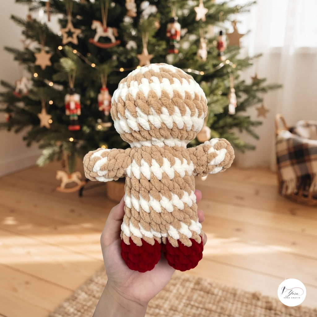 A hand holds a crocheted gingerbread person doll in front of a decorated Christmas tree in a warm, festive room. The doll has a beige and white striped body and hat with red shoes.