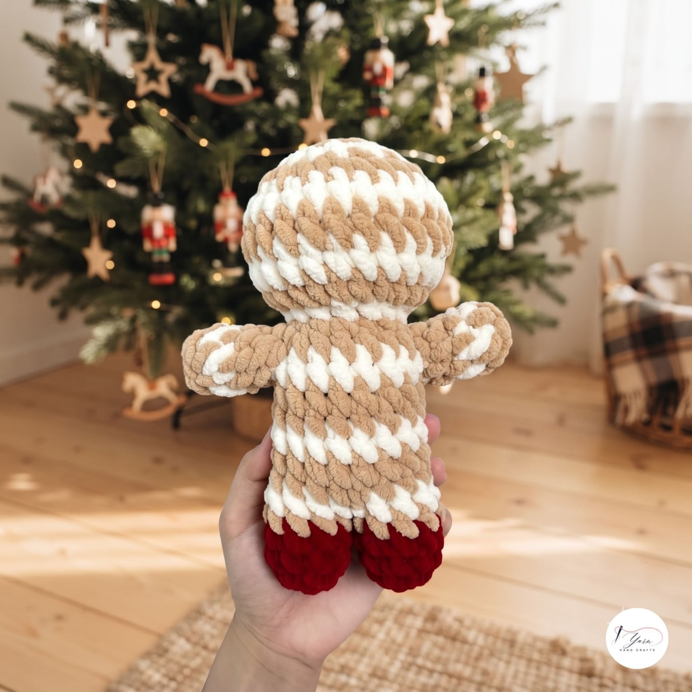 A hand holds a crocheted gingerbread person doll in front of a decorated Christmas tree in a warm, festive room. The doll has a beige and white striped body and hat with red shoes.