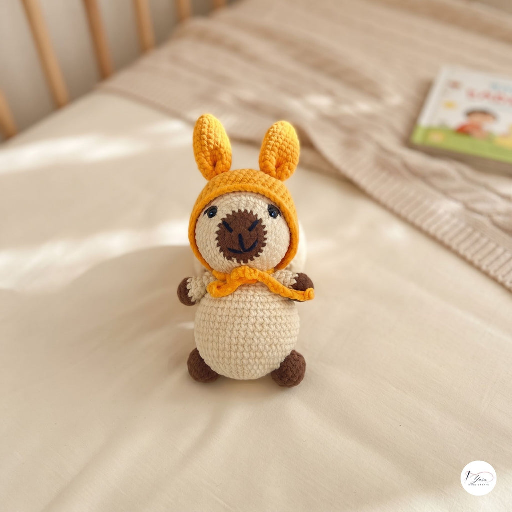 A crocheted llama doll wearing a yellow hood and matching scarf, standing on a cream-colored bed with a wooden headboard in the background.