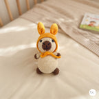 A crocheted llama doll wearing a yellow hood and matching scarf, standing on a cream-colored bed with a wooden headboard in the background.