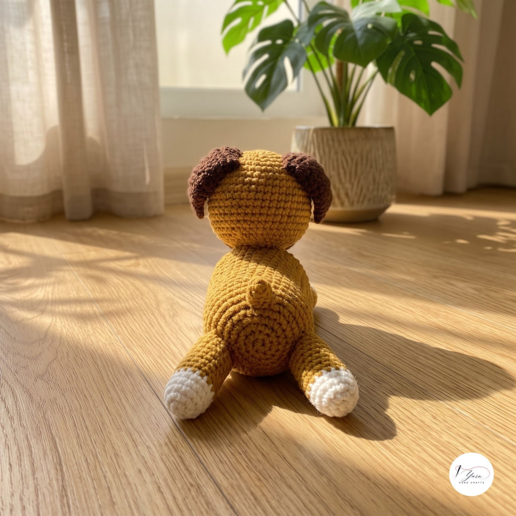 A crocheted plush dog toy sits on a wooden floor in a sunlit room, with sheer curtains, a potted plant, and natural shadows in the background. The toy is mustard yellow with dark brown ears and white paws and tail tip.