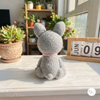 A gray crocheted bunny plush toy with long ears sits on a white wooden table, viewed from behind. The bunny has a small pink collar and is set in a bright, sunlit room with potted plants and a wooden calendar in the background.