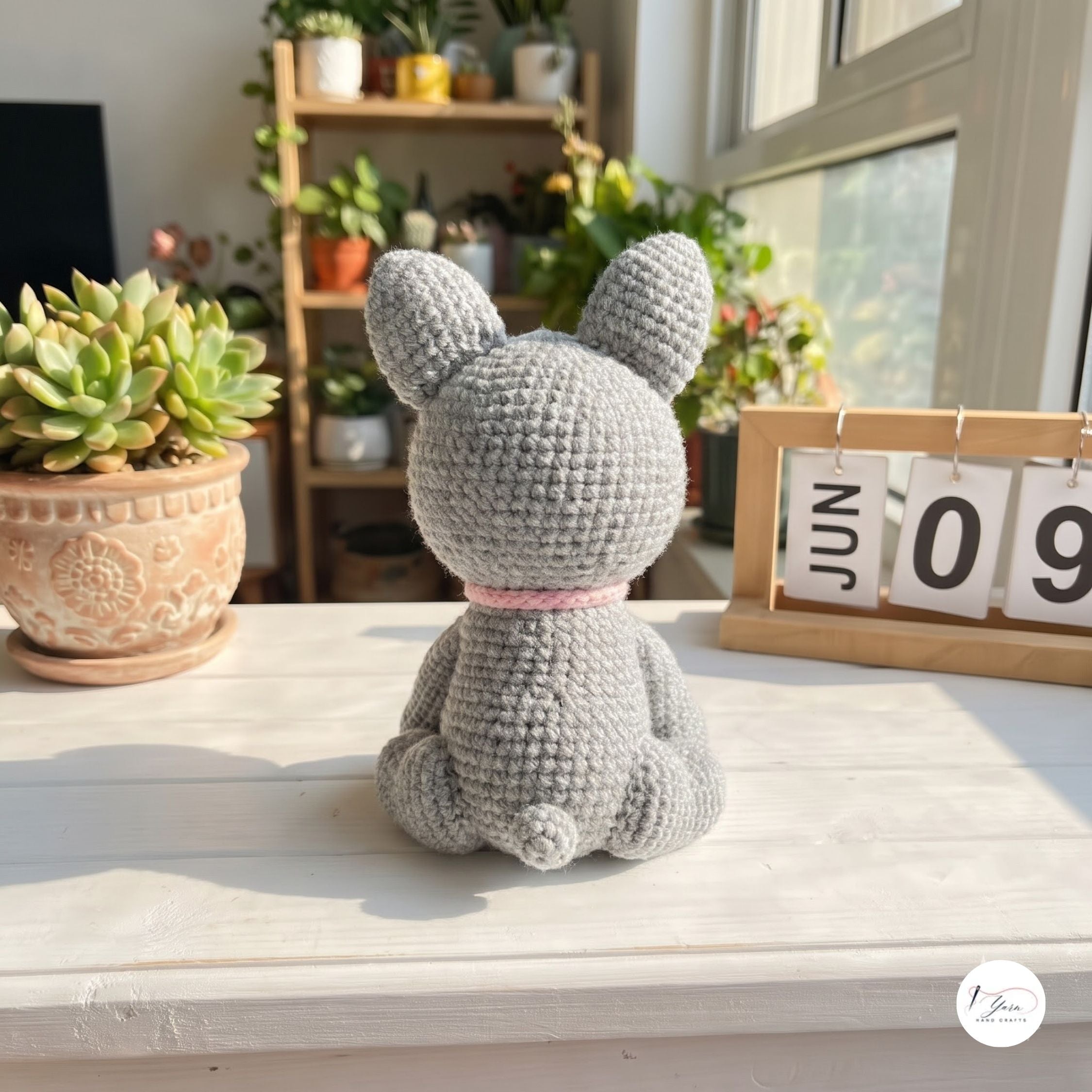 A gray crocheted bunny plush toy with long ears sits on a white wooden table, viewed from behind. The bunny has a small pink collar and is set in a bright, sunlit room with potted plants and a wooden calendar in the background.