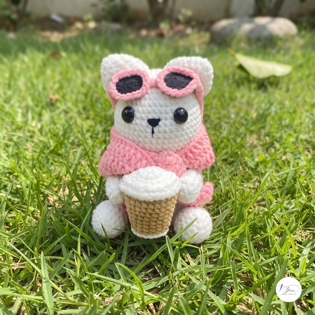 A cute handmade white crochet cat plush sitting outdoors on green grass, wearing a soft pink sweater and pink-framed sunglasses pushed up on its head, holding a crocheted takeaway coffee cup.
