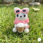 A cute handmade white crochet cat plush sitting outdoors on green grass, wearing a soft pink sweater and pink-framed sunglasses pushed up on its head, holding a crocheted takeaway coffee cup.