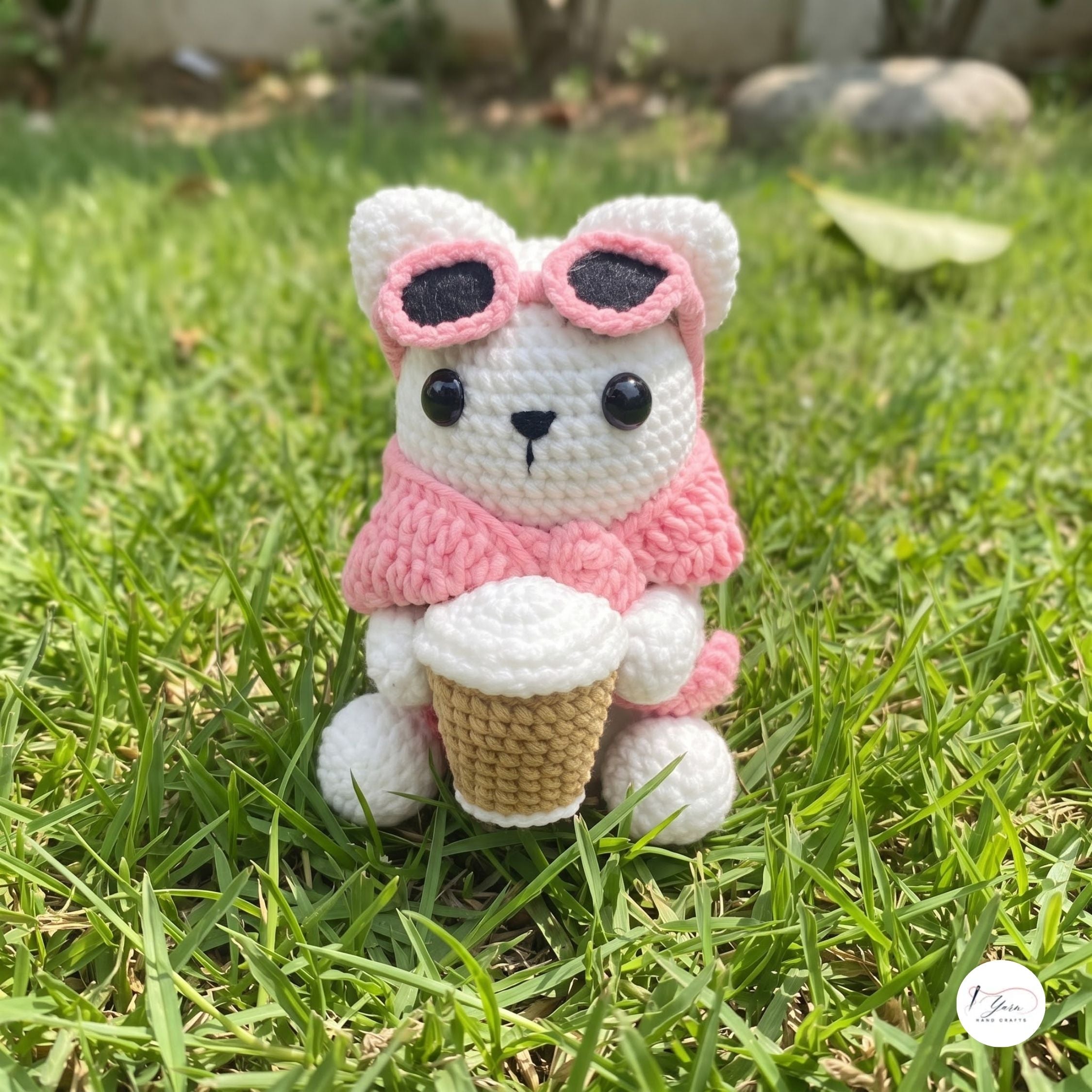 A cute handmade white crochet cat plush sitting outdoors on green grass, wearing a soft pink sweater and pink-framed sunglasses pushed up on its head, holding a crocheted takeaway coffee cup.