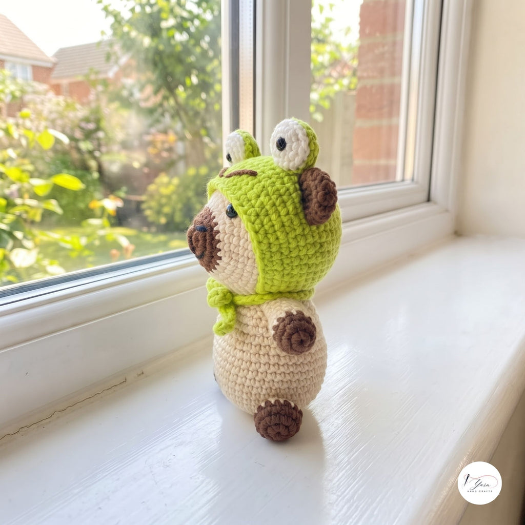 A crocheted plush toy shaped like a small frog or animal character, standing on a white windowsill with a real outdoor garden and brick wall visible behind it. The toy is lime green and cream with brown accents.