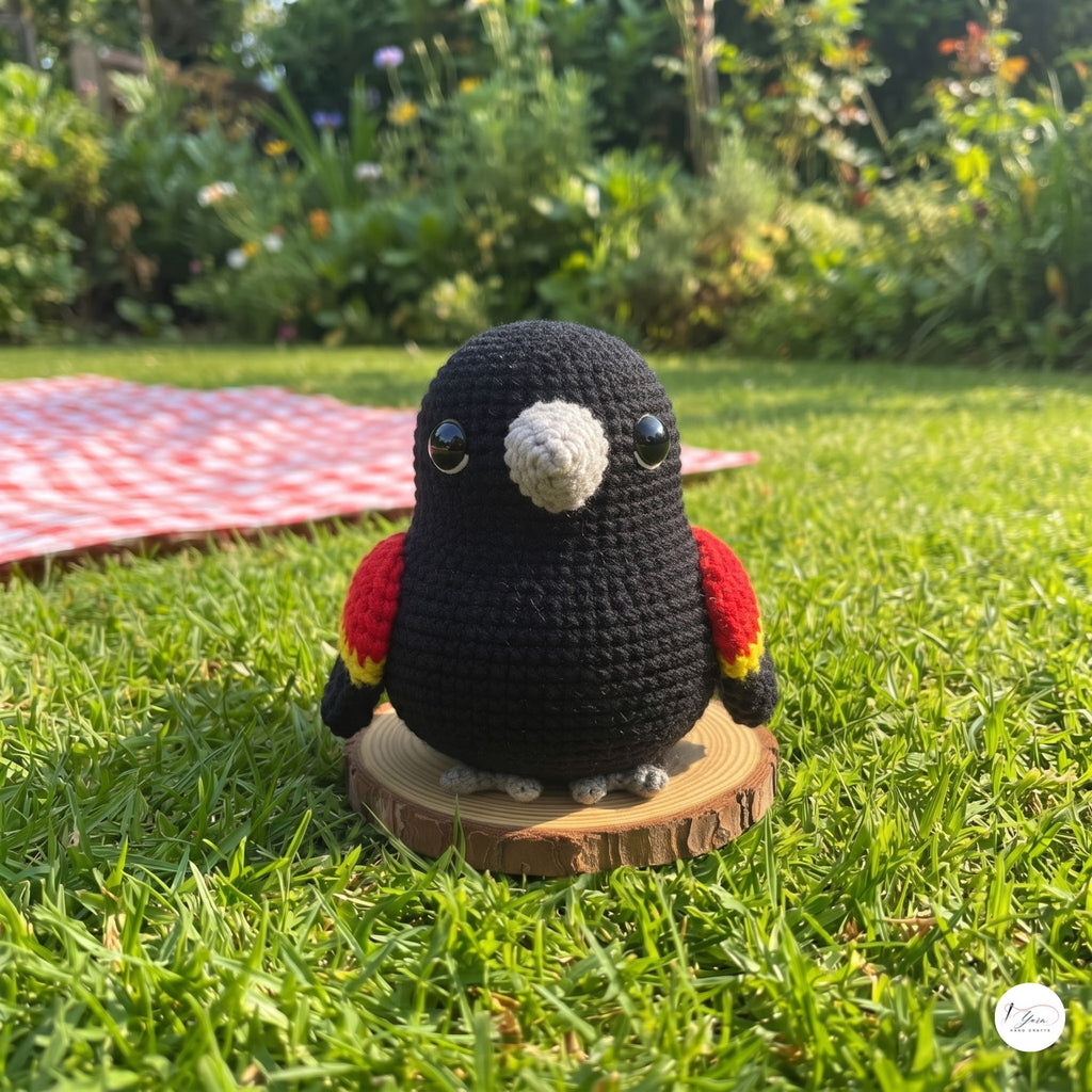 A crocheted black bird toy with red and yellow wings, a white beak, and gray feet, standing on a small round wooden slice in a grassy garden setting.