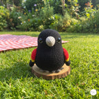 A crocheted black bird toy with red and yellow wings, a white beak, and gray feet, standing on a small round wooden slice in a grassy garden setting.