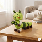 A crocheted plush caterpillar toy sits on a light wood table in a softly lit living room. The toy is bright green with darker green stripes, white accents, and dark brown feet and antennae.