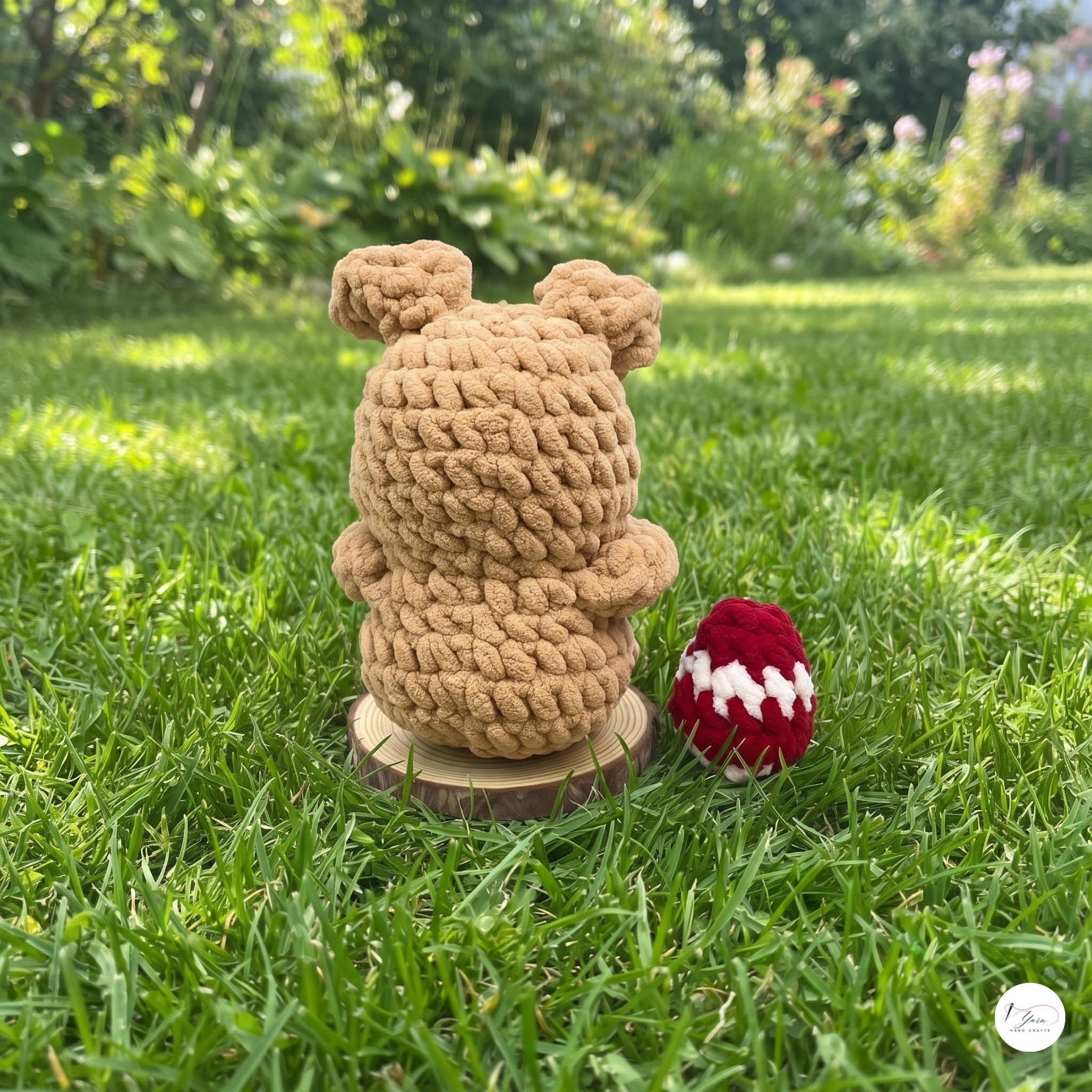 A tan crocheted bear figurine sitting on a small wooden base next to a red and white crocheted egg, placed on a green grass lawn with a blurred garden background.
