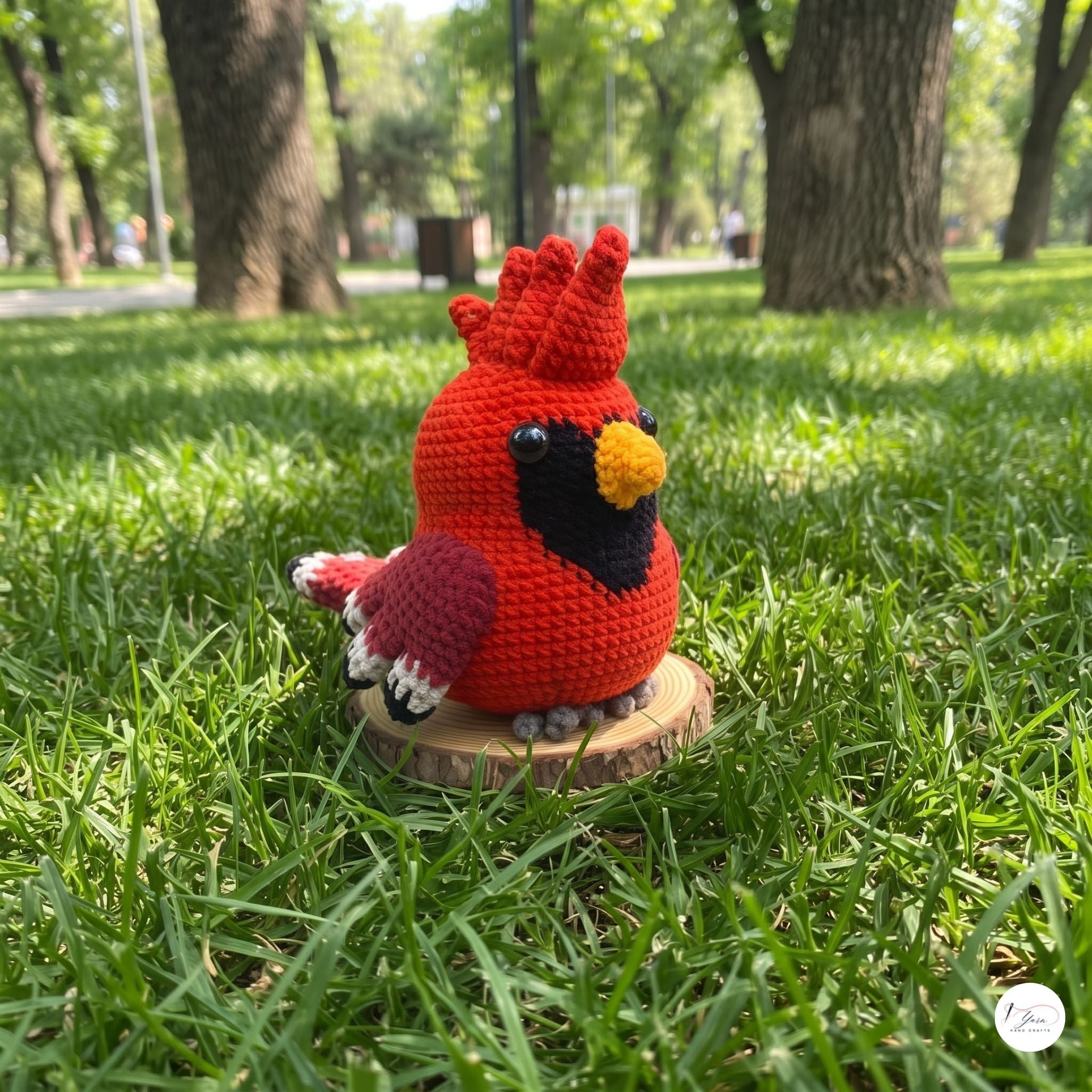 A red crocheted cardinal bird toy sits on a small round wooden slice in a grassy park with trees in the background.