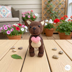 A crocheted brown bear toy standing on a wooden table, holding a pink heart. The background shows a cozy outdoor patio setting with potted flowers, a wicker chair, and a wooden trellis.
