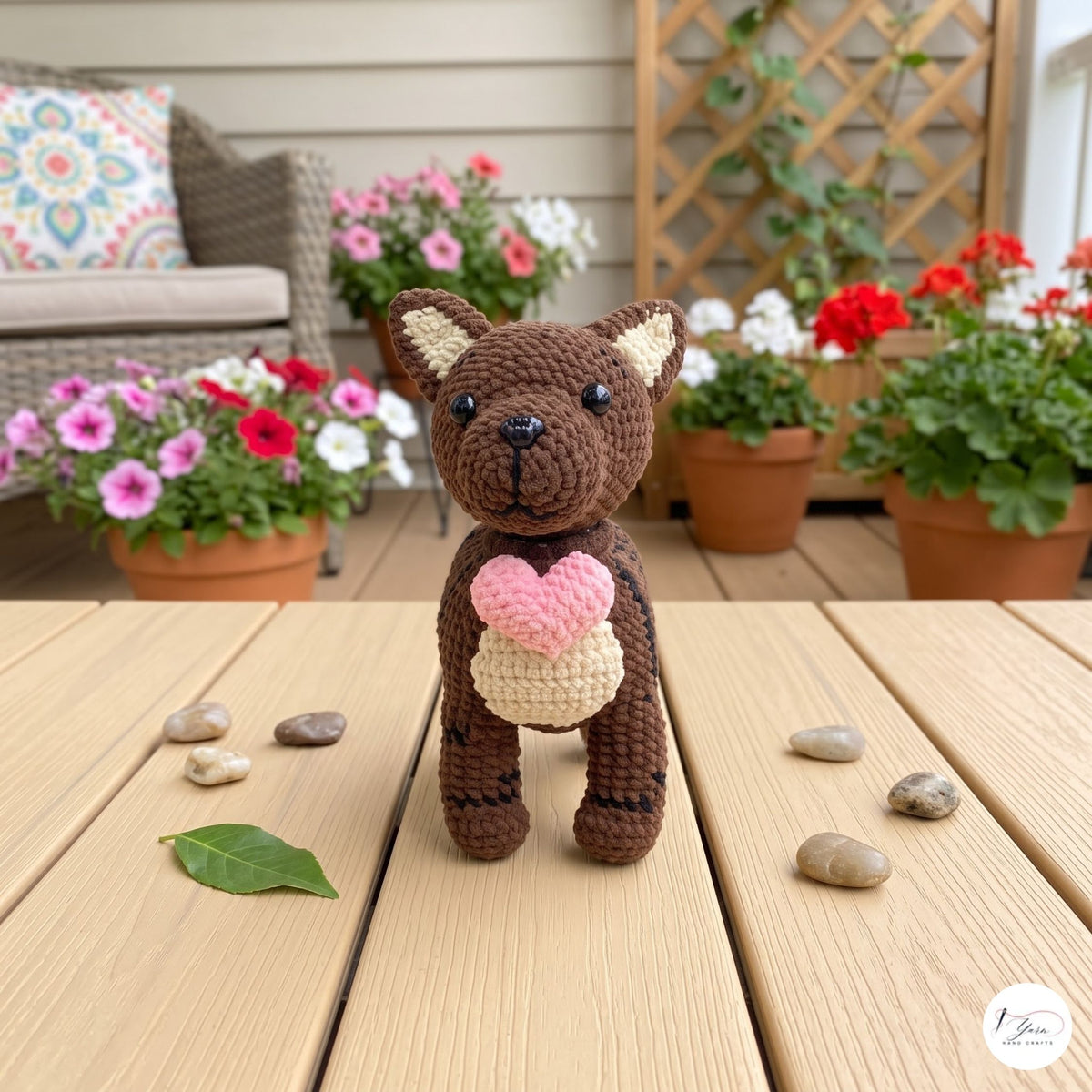 A crocheted brown bear toy standing on a wooden table, holding a pink heart. The background shows a cozy outdoor patio setting with potted flowers, a wicker chair, and a wooden trellis.