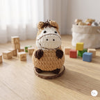 A crocheted plush toy horse standing on a round wooden base, placed on a light wood tabletop with colorful toy blocks and a soft fabric basket in the background.