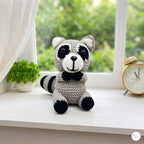 A crocheted raccoon plush toy sits on a white windowsill. The toy is made of light gray and black yarn with a white snout and nose.
