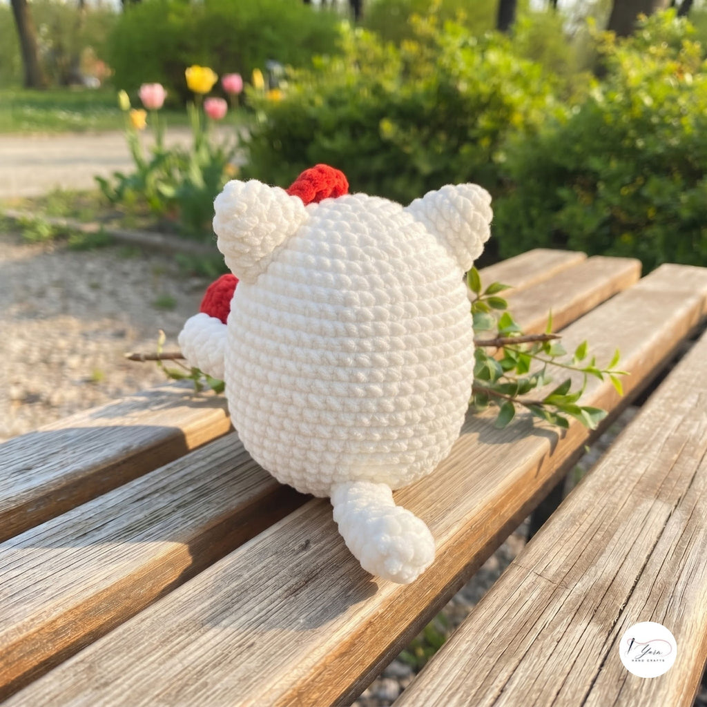 A crocheted white bear plush toy with red accents sits on a weathered wooden bench in a sunny park setting with green bushes and flowers in the background.