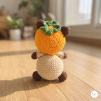 A crocheted plush toy shaped like a persimmon with a cream-colored body, brown limbs, and a green leafy top, sitting on a wooden floor in a softly blurred indoor setting.