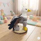 A crocheted eagle toy standing on a round wood slice base in a softly lit children's room. The toy has a white head and neck, gray body, dark navy and brown wings, and yellow feet and beak.