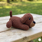A crocheted plush puppy toy in medium brown lies on a light wooden table outdoors, with a blurred green grassy background.