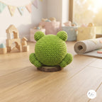 A green crocheted frog toy sits on a small round wooden base on a light wood floor, with a softly blurred nursery-like background containing toys and pastel decor.