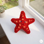 A red crocheted starfish toy with orange spots and small blue eyes sits on a white windowsill with natural light.