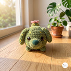 A crocheted plush dog toy in olive green yarn with small yellow speckles, a dark green nose, and a small pink flower on its head, sitting on a wooden windowsill with warm sunlight and potted plants in the background.
