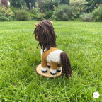 A crocheted toy horse with a brown mane and tail, standing on a round wooden base in a grassy garden.