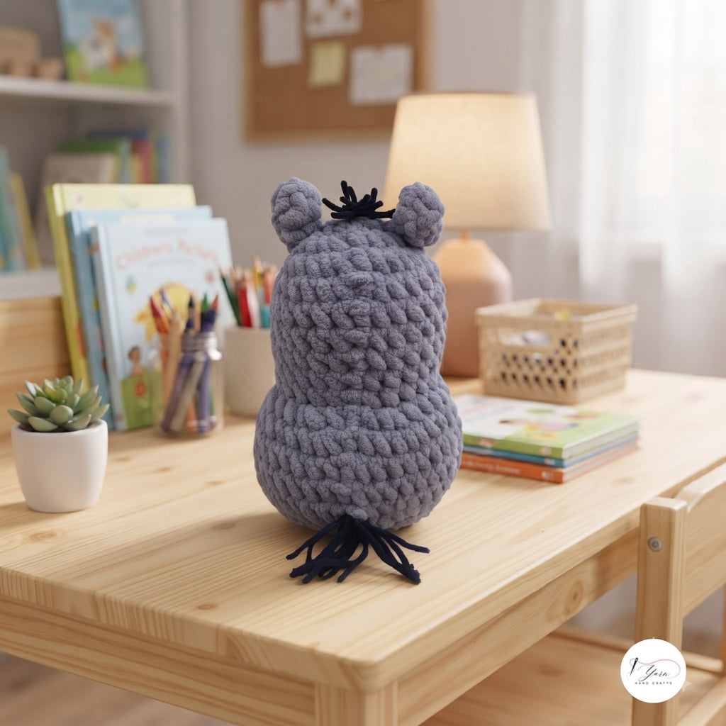 A gray crocheted monster plush toy with black antlers and a black tail sits on a light wood desk in a cozy, softly lit room with books, a lamp, and a small plant in the background.