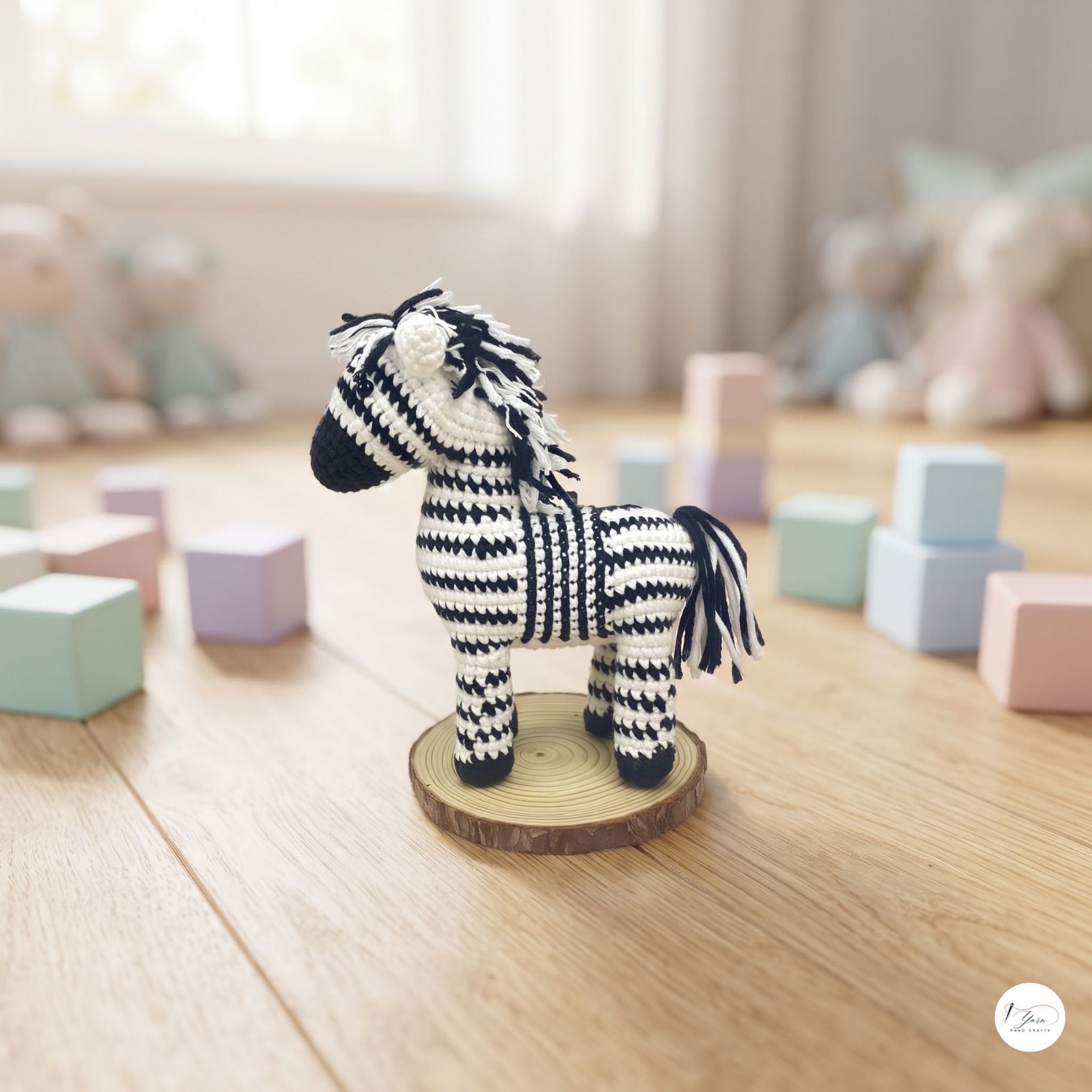 A small crocheted zebra doll with black and white stripes stands on a round wooden slice base on a light wood floor, surrounded by pastel toy blocks and soft-focus stuffed animals in the background.
