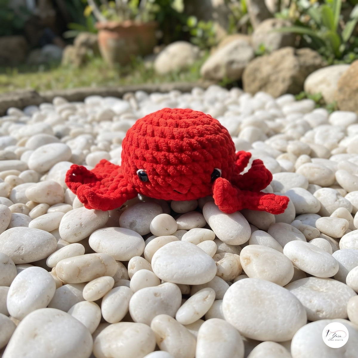 A red crocheted crab toy with small black eyes, resting on a bed of smooth white pebbles outdoors with blurred rocks and greenery in the background.