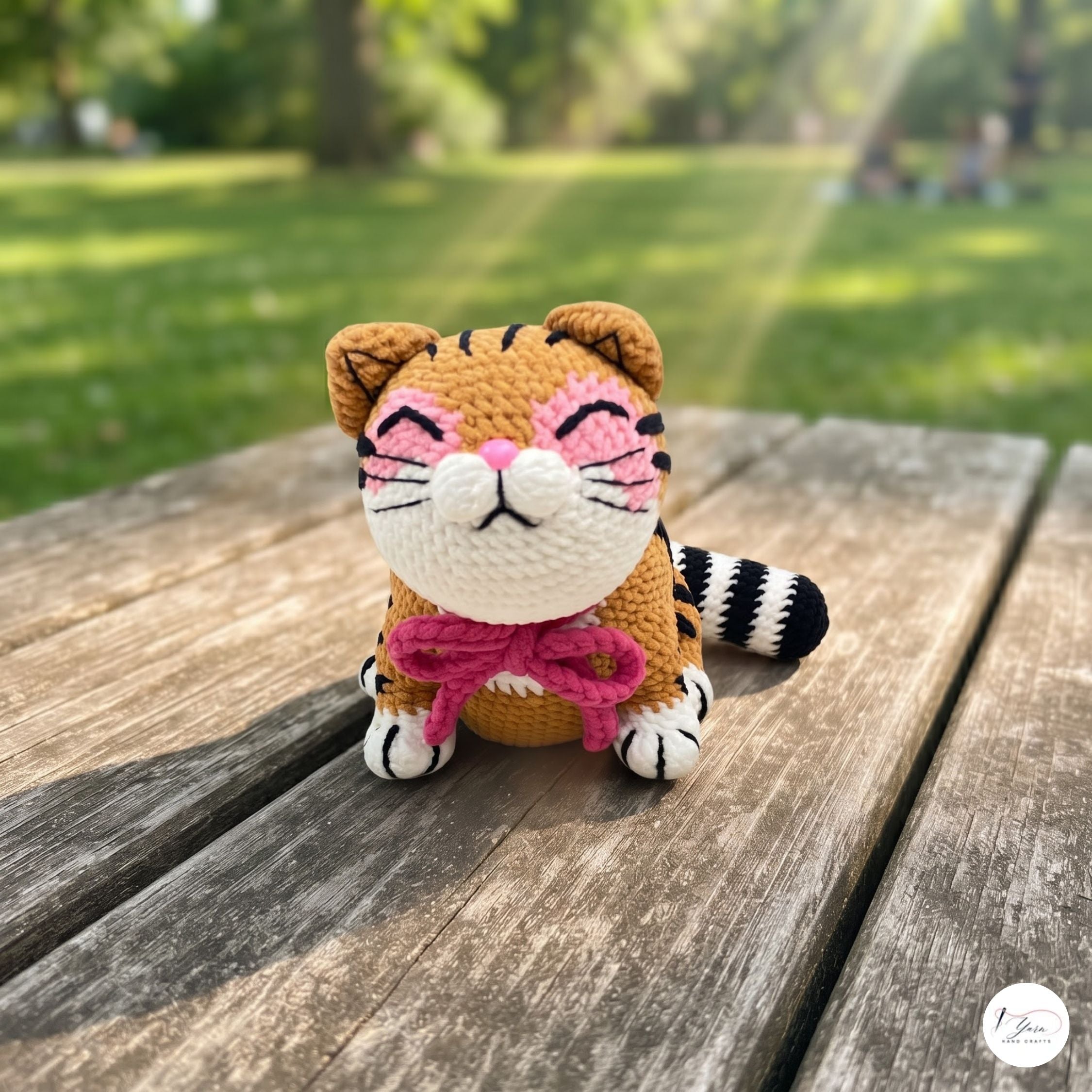 A crocheted tiger toy sits on a weathered wooden table in a sunny park. The toy features orange and black stripes, a cream-colored face and paws, pink stitched cheeks and a pink bow around its neck, with a blurred green park background.