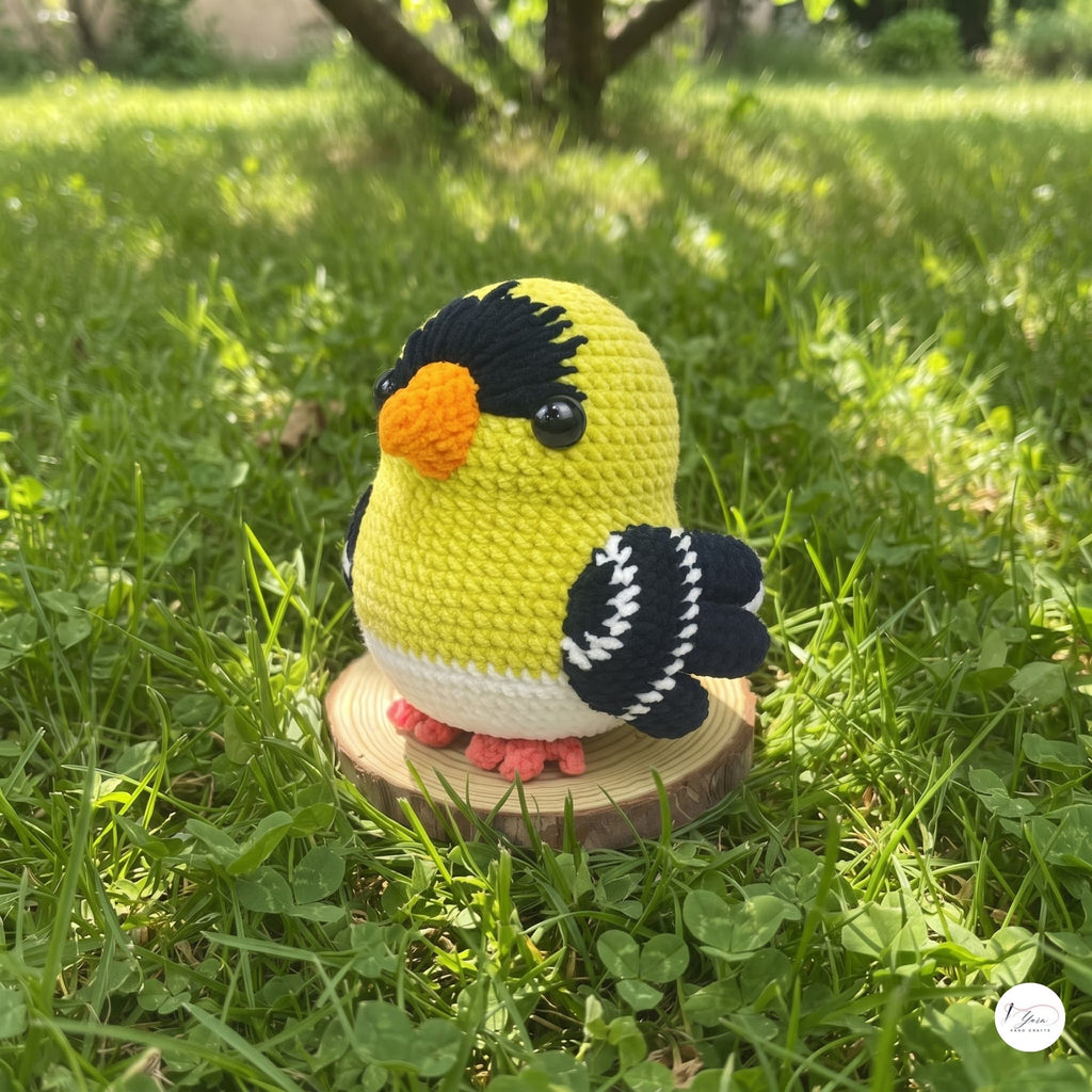 A crocheted goldfinch toy bird sits on a small round wooden slice placed in green grass outdoors. The bird is yellow with black head and wing details, a white belly, an orange beak, and pink feet.