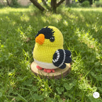 A crocheted goldfinch toy bird sits on a small round wooden slice placed in green grass outdoors. The bird is yellow with black head and wing details, a white belly, an orange beak, and pink feet.