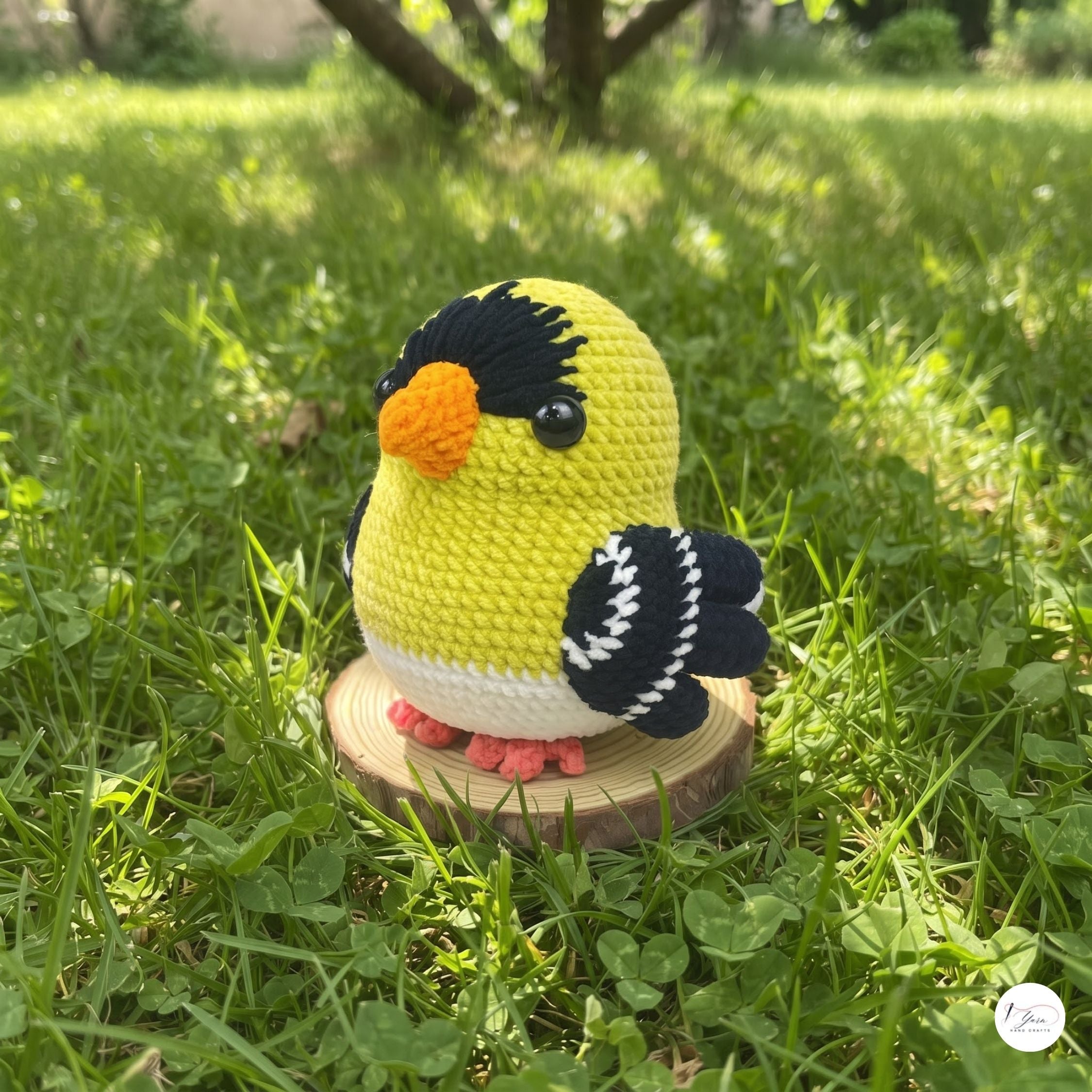 A crocheted goldfinch toy bird sits on a small round wooden slice placed in green grass outdoors. The bird is yellow with black head and wing details, a white belly, an orange beak, and pink feet.