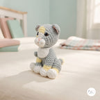 A small crocheted cat toy sits on a light cream bed in a softly lit room. The toy is gray with white and pale yellow accents, including a white chest, paws, and face, and yellow patches on the forehead and hind legs.