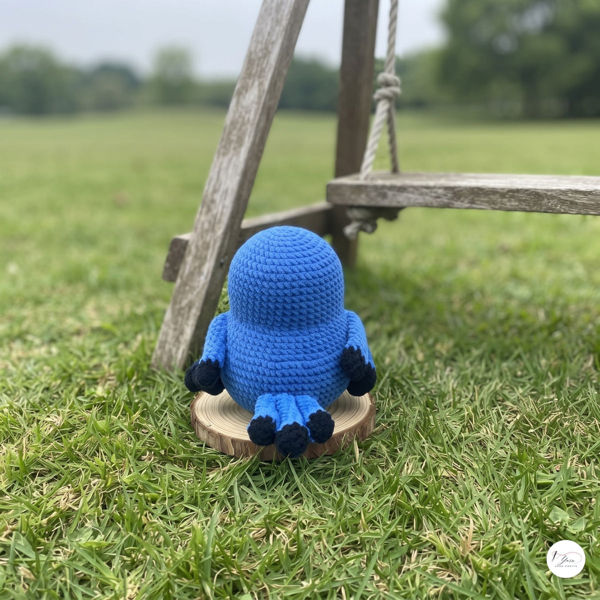 A small crocheted blue amigurumi character with black feet sits on a round wooden slice on a grassy lawn, with a rustic wooden swing leg in the background.