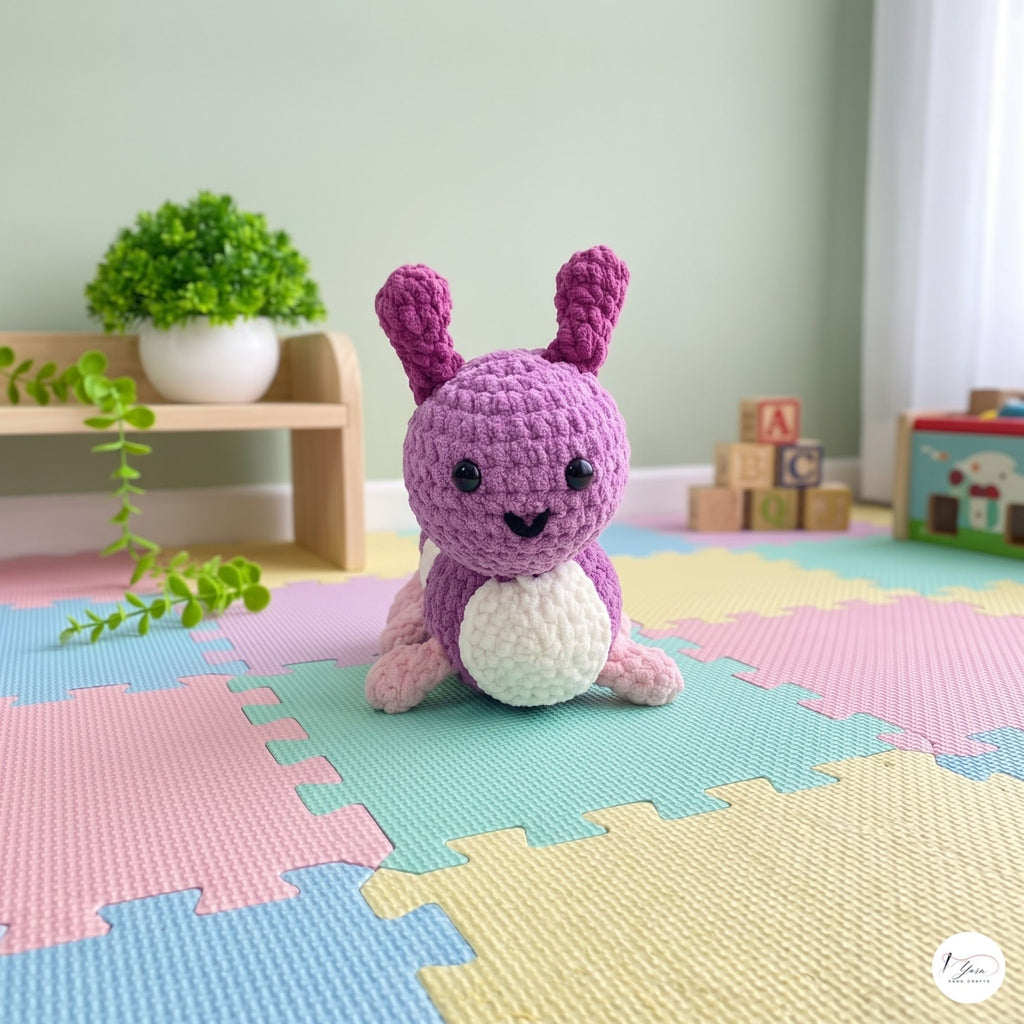 A purple and white crocheted bunny toy sits on a pastel interlocking foam play mat in a softly styled children's room. The background includes a small wooden shelf with a potted plant, colorful toy blocks, and a small toy car.