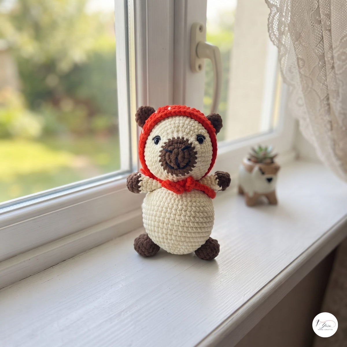 A crocheted plush bear toy with a cream body, brown limbs and snout, wearing a red hood, sitting on a white windowsill with a blurred outdoor garden background.