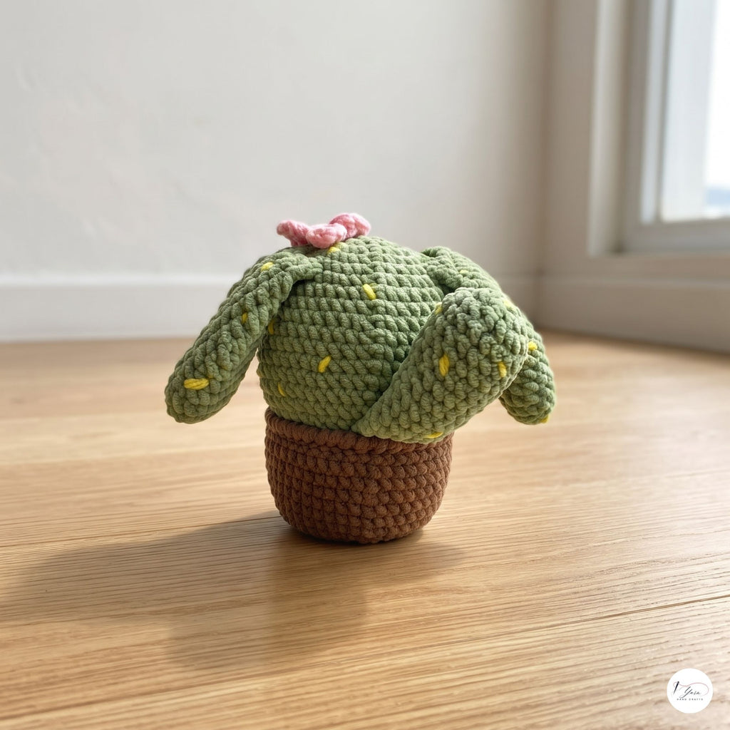 A plush handmade crochet cactus puppy toy with long floppy ears, yellow embroidered spines, and a pink flower on top, sitting in a brown crochet pot base on a sunlit wooden floor.