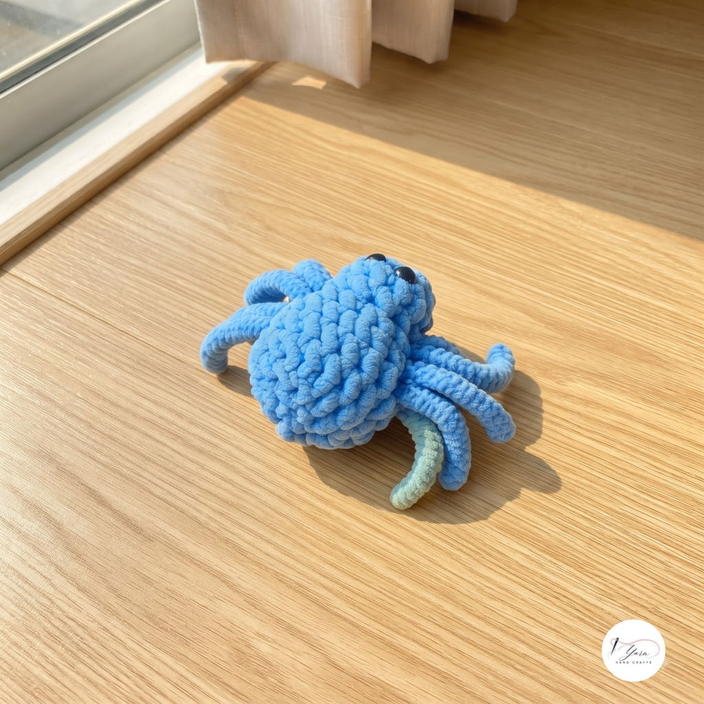 A close-up view of a plush handmade crochet spider toy in light blue with glossy black safety eyes, sitting on a light wooden floor under bright natural sunlight.