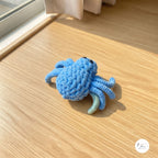 A close-up view of a plush handmade crochet spider toy in light blue with glossy black safety eyes, sitting on a light wooden floor under bright natural sunlight.