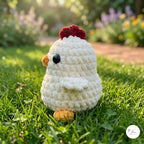 A small crocheted chicken toy sits on green grass outdoors with a blurred garden background. The toy is primarily off-white with a dark red comb, a small orange beak and feet, and a black bead eye.