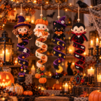 Four handmade crocheted Halloween hanging ornaments displayed on a rustic branch in a cozy, warmly lit holiday setting. The ornaments include a witch, a pumpkin, a black cat, and a vampire, each with detailed stitched features and themed accessories like brooms, cauldrons, bats, and coffins.