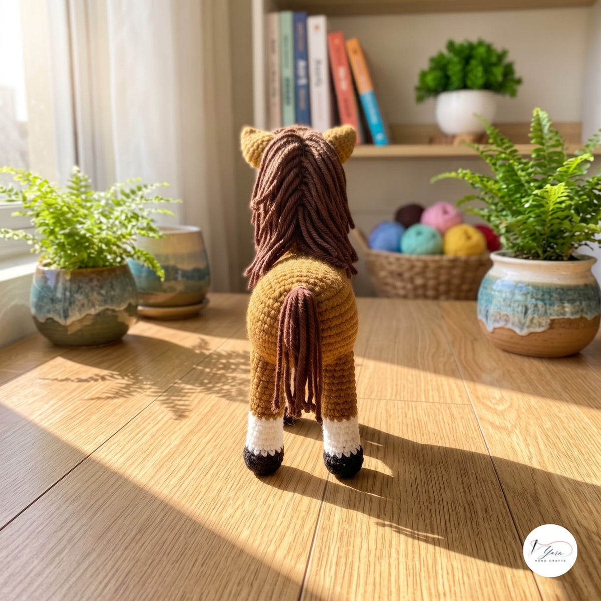 A crocheted plush horse toy shown from the back on a wooden table in a sunlit room, with potted ferns, books, and yarn balls in the background.