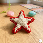 A crocheted starfish toy in red and cream, placed on a light wood floor with seashell props and a colorful rug in the background.