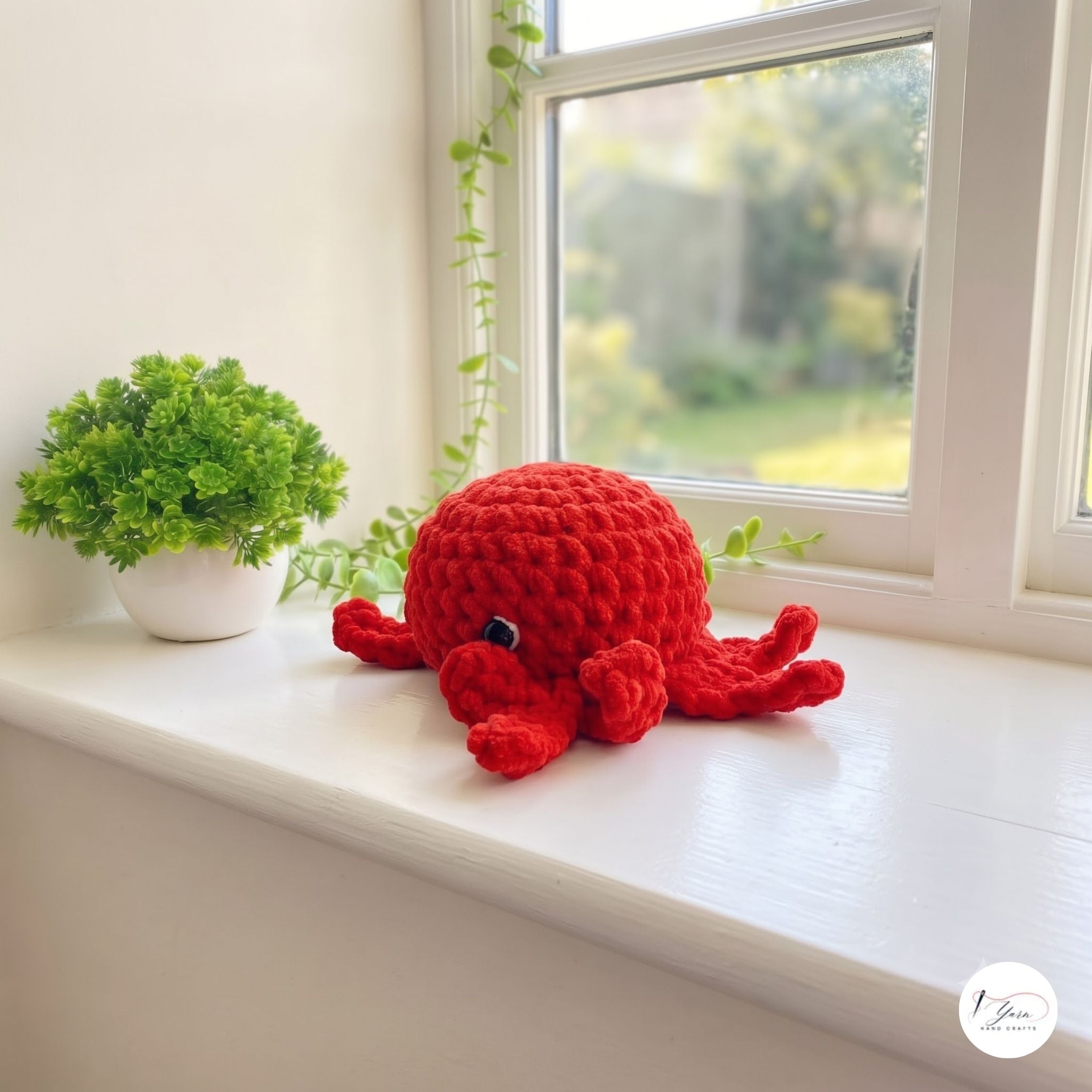 A red crocheted octopus plush toy sits on a white windowsill next to a small potted green plant, with a blurred outdoor garden view in the background.