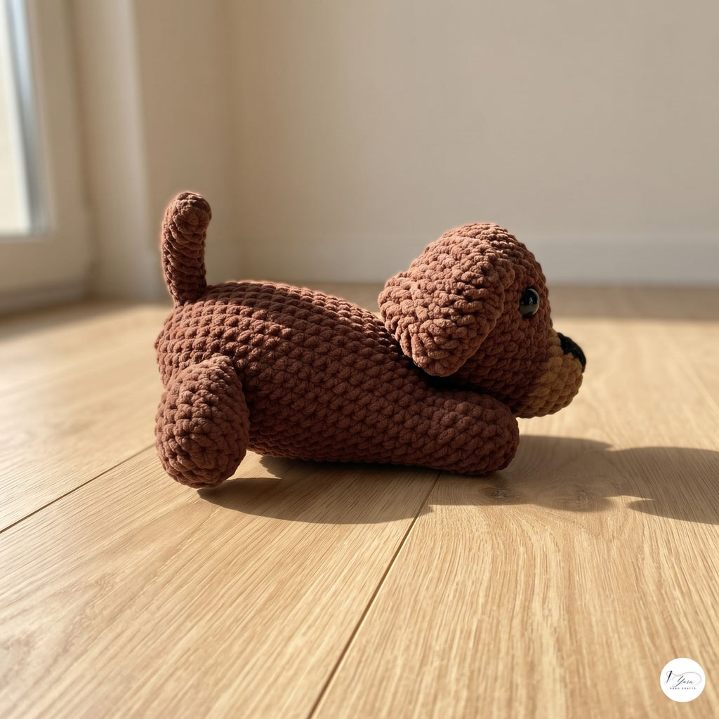 A brown crocheted plush dog toy lying on a light wood floor in a sunlit room.
